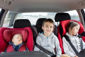 Drei Geschwister sitzen nebeneinander in schmalen Kindersitzen im Auto.
